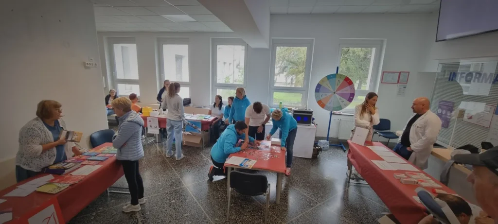 Na sliki je notranjost zdravstvene ustanove, kjer se odvija dogodek s promocijo zdravja – ljudje stojijo ob mizah z informativnimi materiali, sodelujejo v aktivnostih in se pogovarjajo z zdravstvenimi delavci.