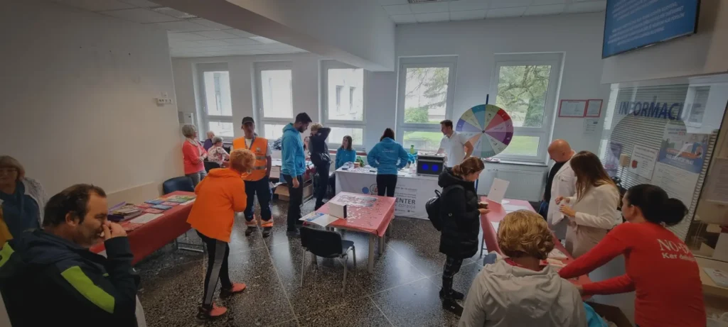 Na sliki je notranjost zdravstvene ustanove, kjer se odvija dogodek s promocijo zdravja – ljudje stojijo ob mizah z informativnimi materiali, sodelujejo v aktivnostih in se pogovarjajo z zdravstvenimi delavci.