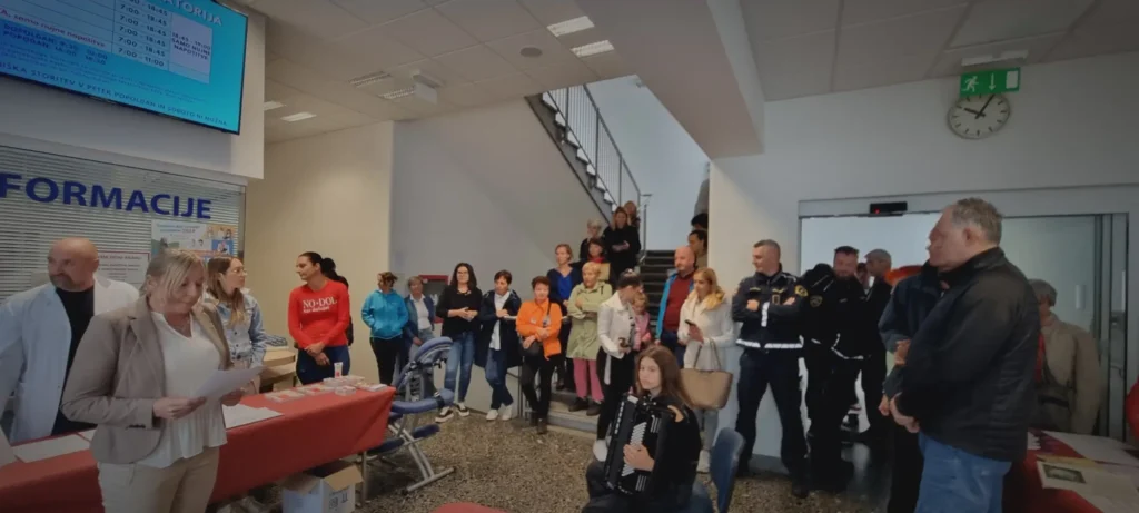 Na sliki je notranjost zdravstvene ustanove, kjer se odvija dogodek s promocijo zdravja – ljudje stojijo ob mizah z informativnimi materiali, sodelujejo v aktivnostih in se pogovarjajo z zdravstvenimi delavci.