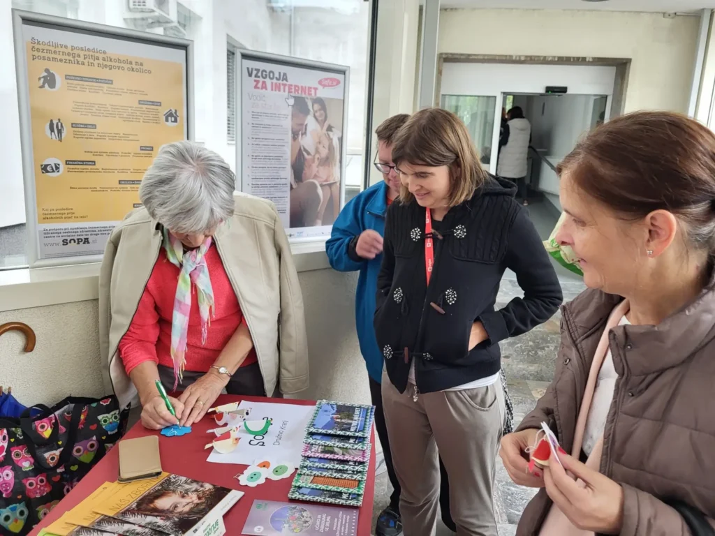 Na sliki je notranjost zdravstvene ustanove, kjer se odvija dogodek s promocijo zdravja – ljudje stojijo ob mizah z informativnimi materiali, sodelujejo v aktivnostih in se pogovarjajo z zdravstvenimi delavci.