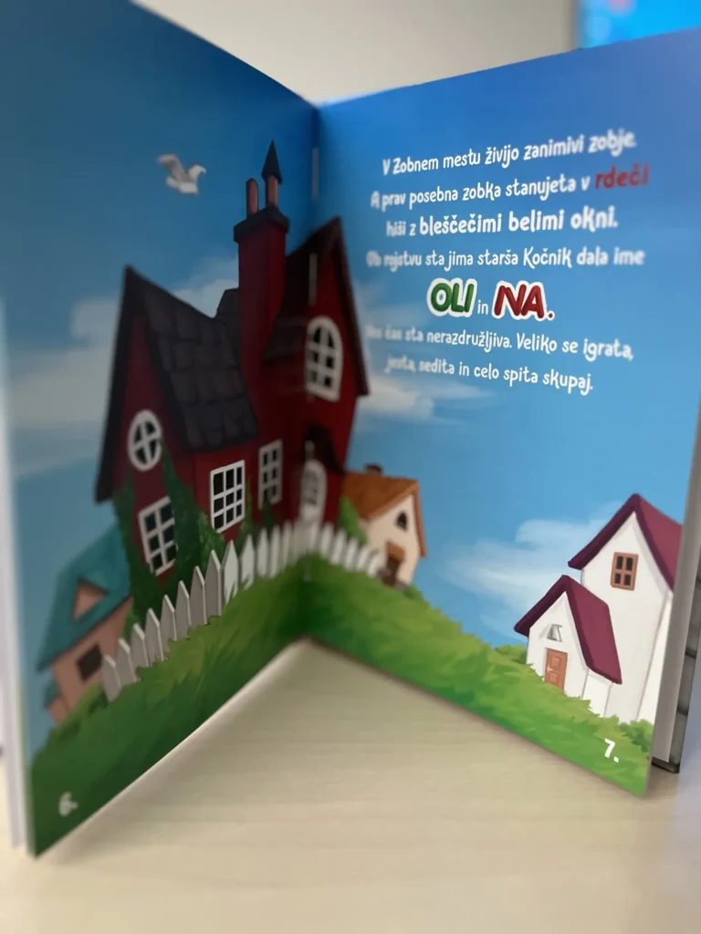 Na sliki je ilustrirana notranja stran otroške knjige, kjer je na desni strani besedilo o zobkih Oliju in Na, ki živita v rdeči hiši z belimi okni v Zobnem mestu, na levi pa ilustracija pisanih hiš z ograjo in zelenico.